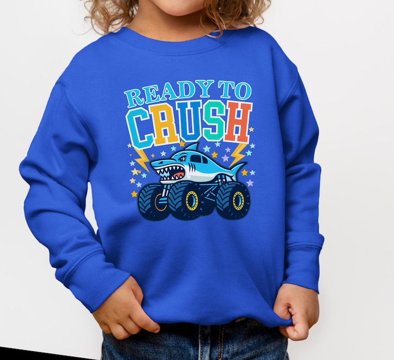 Pu&ograve; includere: Una felpa blu con una grafica di un monster truck a forma di squalo e la scritta "READY TO CRUSH". Lo squalo &egrave; azzurro e bianco. Il testo &egrave; in arancione, verde e blu.