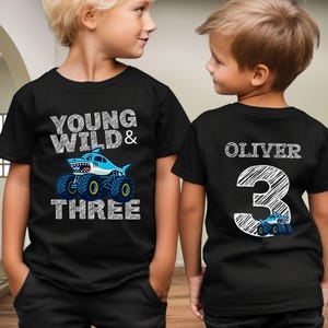 Op de afbeelding: Zwarte T-shirts met een verjaardagsthema. De voorkant van het shirt zegt "Young Wild & Three" met een haaien-monster truck-afbeelding. De achterkant van het shirt heeft de naam "Oliver" en het nummer "3" met een monstertruck.