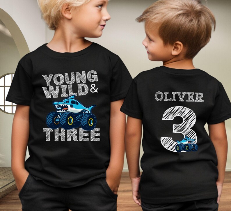 Op de afbeelding: Zwarte T-shirts met een verjaardagsthema. De voorkant van het shirt zegt "Young Wild & Three" met een haaien-monster truck-afbeelding. De achterkant van het shirt heeft de naam "Oliver" en het nummer "3" met een monstertruck.
