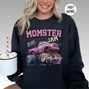 Pu&ograve; includere: Felpa nera con una grafica di un monster truck rosa e il testo "Momster Jam Mommy".