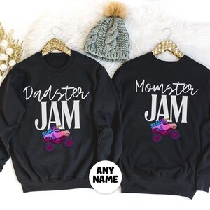 May include: Black sweatshirts with the phrases "Dadster JAM" and "Momster JAM" in white script. Each shirt features a colorful monster truck with a unicorn horn. A gray knit beanie with a pom-pom is also visible.