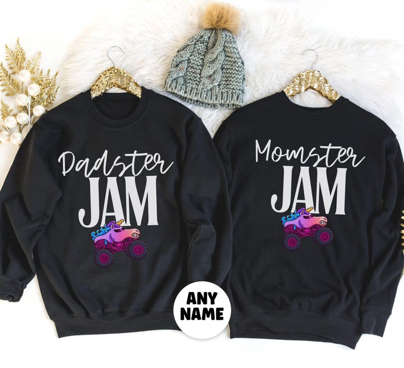 May include: Black sweatshirts with the phrases "Dadster JAM" and "Momster JAM" in white script. Each shirt features a colorful monster truck with a unicorn horn. A gray knit beanie with a pom-pom is also visible.