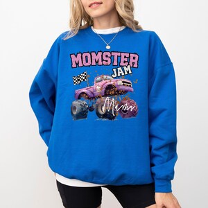 Pu&ograve; includere: Una felpa blu reale con una grafica di un monster truck rosa e il testo "Momster Jam" in rosa. Il camion ha una bandiera a scacchi e la parola "Mom" &egrave; scritta in rosa sul lato.