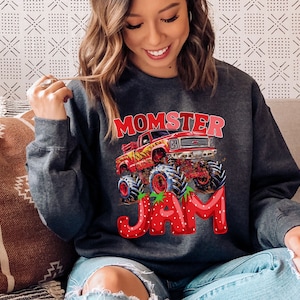 Pictured is a woman wearing a dark gray sweatshirt with a red monster truck graphic and the text "Momster Jam" in white and red letters. The word JAM are strawberry letters
