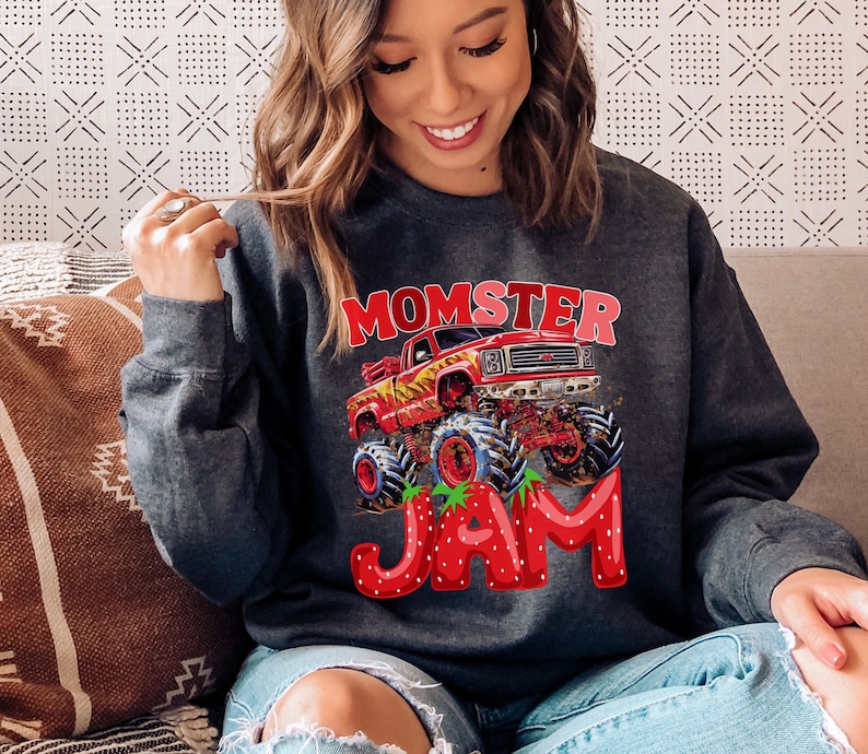 Pictured is a woman wearing a dark gray sweatshirt with a red monster truck graphic and the text "Momster Jam" in white and red letters. The word JAM are strawberry letters