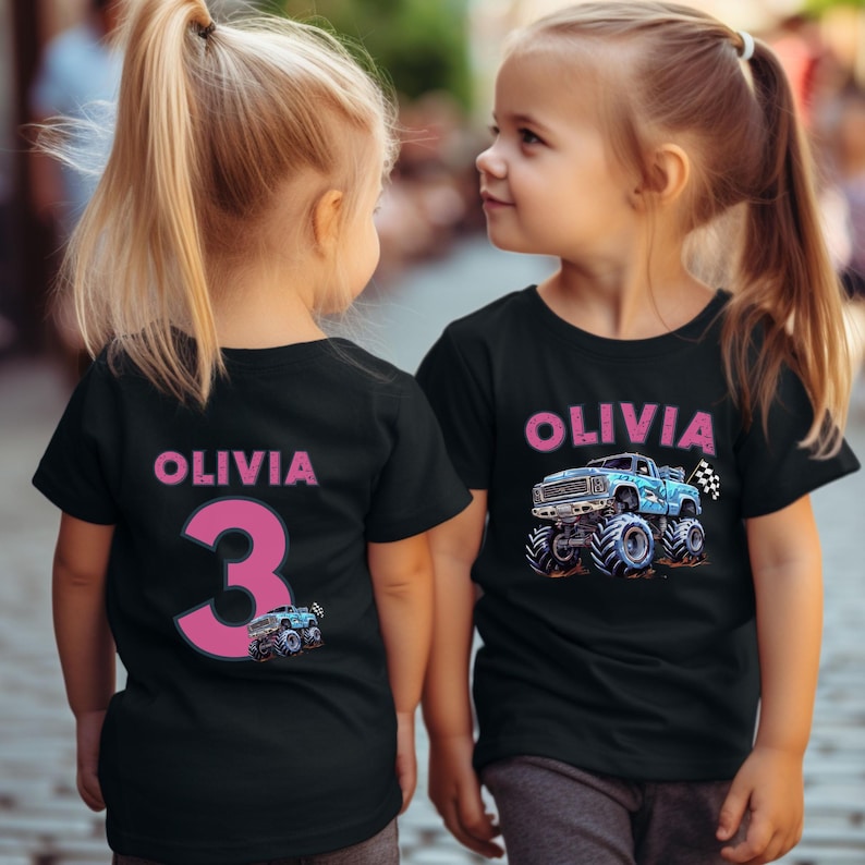 May include: Two young girls wearing matching black t-shirts with the name "OLIVIA" in pink. One shirt has a large pink number "3" and a small monster truck graphic. The other shirt features a blue monster truck graphic and the name "OLIVIA".