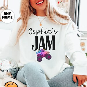May include: White sweatshirt with the text "Sophia's JAM" in black script and block letters. A cartoon monster truck with a unicorn head is below the text. The truck has purple wheels and a pink body.