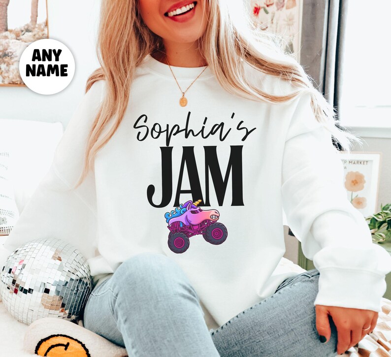 May include: White sweatshirt with the text "Sophia's JAM" in black script and block letters. A cartoon monster truck with a unicorn head is below the text. The truck has purple wheels and a pink body.