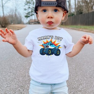 May include: A white t-shirt with the text "MAMA'S BOY" above a cartoon shark on monster truck wheels. The shark is blue and white, with a red and yellow burst background. The shirt is worn by a young child.