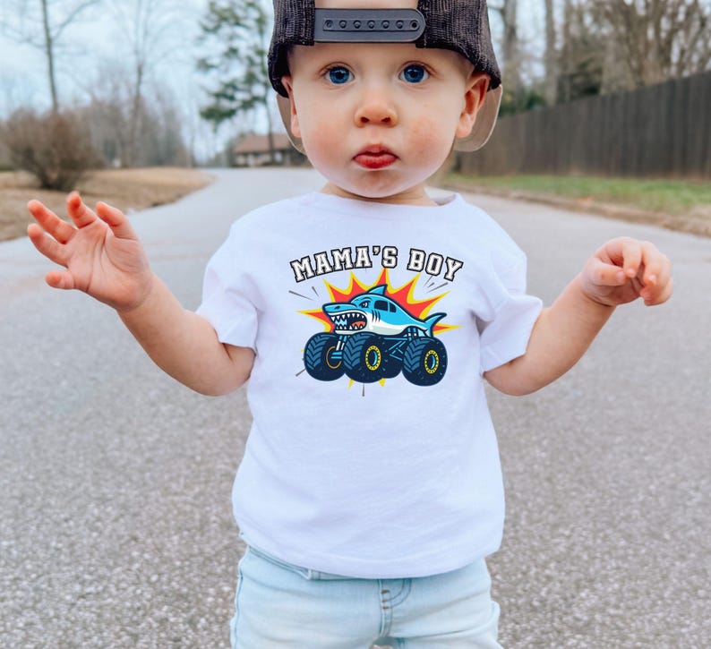 May include: A white t-shirt with the text "MAMA'S BOY" above a cartoon shark on monster truck wheels. The shark is blue and white, with a red and yellow burst background. The shirt is worn by a young child.
