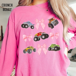 K&ouml;nnte beinhalten: Rosa Langarmshirt mit einem Design aus bunten Monstertrucks und rosa Schleifen. Der Text "CRUNCH BERRY" ist oben links aufgedruckt. Die Monstertrucks sind in verschiedenen Farben, darunter Blau, Orange, Rot, Gelb und Gr&uuml;n.
