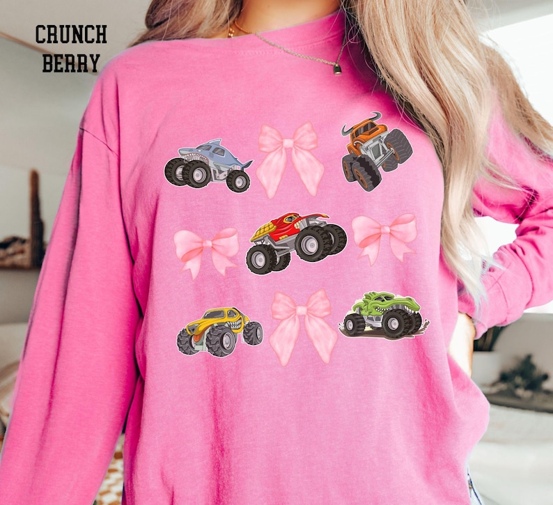 K&ouml;nnte beinhalten: Rosa Langarmshirt mit einem Design aus bunten Monstertrucks und rosa Schleifen. Der Text "CRUNCH BERRY" ist oben links aufgedruckt. Die Monstertrucks sind in verschiedenen Farben, darunter Blau, Orange, Rot, Gelb und Gr&uuml;n.