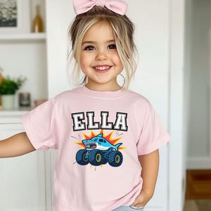 May include: A light pink t-shirt with the name "ELLA" in a bold, black and white font. Below the name is a cartoon shark on a monster truck, with blue wheels and a yellow and red burst background.