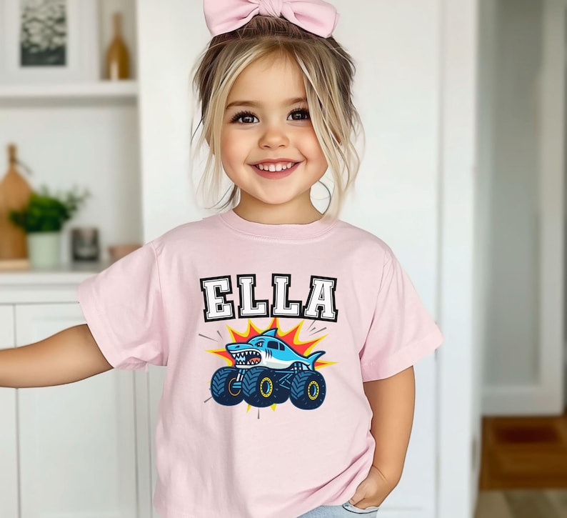 May include: A light pink t-shirt with the name "ELLA" in a bold, black and white font. Below the name is a cartoon shark on a monster truck, with blue wheels and a yellow and red burst background.