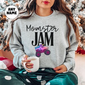 May include: A light gray sweatshirt with the text "Momster JAM" in black script and block letters. A colorful monster truck with a unicorn head is below the text. The sweatshirt has long sleeves.