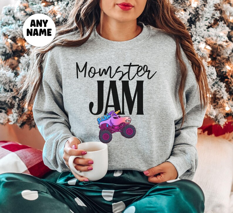 May include: A light gray sweatshirt with the text "Momster JAM" in black script and block letters. A colorful monster truck with a unicorn head is below the text. The sweatshirt has long sleeves.