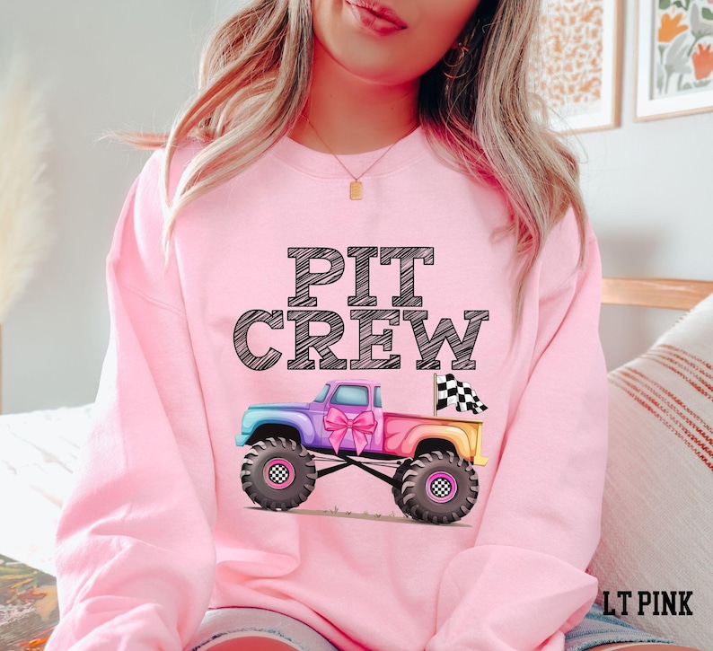 model is wearing a pretty pink monster truck sweatshirt with watercolor rainbow colors and coquette  black and white checkered flag sketch big graphic letters family matching different version for male and female pink for the girls and purple/blue for the guys