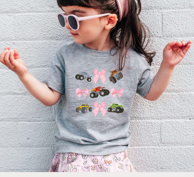 Op de afbeelding: Een grijs t-shirt met kleurrijke monster truck graphics en roze strikken. Het shirt wordt gedragen door een kind, gecombineerd met een bloemenrok en een roze zonnebril. Het ontwerp bevat verschillende kleuren monster trucks.