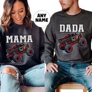 以下が含まれることがあります： 「MAMA」と「DADA」の白いブロック文字がプリントされた、2枚の濃いグレーのスウェットシャツ。それぞれ、黒と赤のモンスターカーのグラフィックが特徴で、スカルと炎が描かれています。スウェットシャツは青いジーンズと合わせて着用されています。