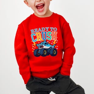 Pu&ograve; includere: Una felpa rossa con la scritta "READY TO CRUSH" in lettere colorate. La grafica presenta uno squalo alla guida di un monster truck con ruote blu e nere. Il design include stelle e fulmini.