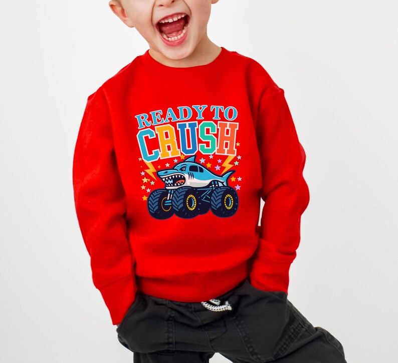 Pu&ograve; includere: Una felpa rossa con la scritta "READY TO CRUSH" in lettere colorate. La grafica presenta uno squalo alla guida di un monster truck con ruote blu e nere. Il design include stelle e fulmini.