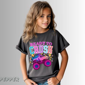 May include: Dark gray t-shirt with the text "READY TO CRUSH" in pink and blue. The shirt features a colorful graphic of a unicorn monster truck with pink wheels and a purple body. The shirt is worn by a young person.