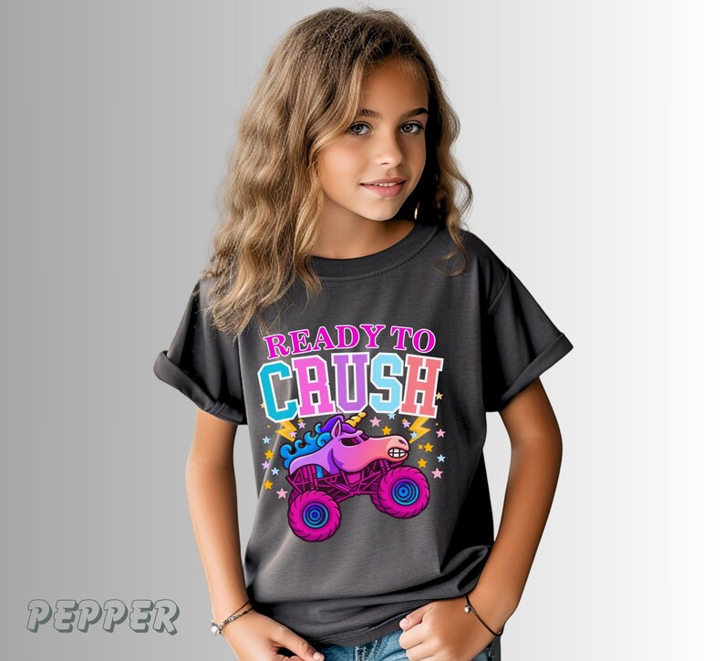 May include: Dark gray t-shirt with the text "READY TO CRUSH" in pink and blue. The shirt features a colorful graphic of a unicorn monster truck with pink wheels and a purple body. The shirt is worn by a young person.