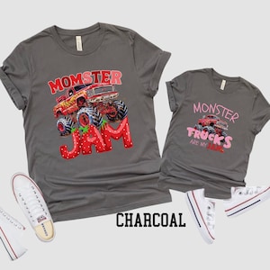 Two charcoal gray t-shirts with monster truck graphics. One shirt says "Momster Jam" with a red monster truck and strawberry design. The other says "Monster Trucks are my Jam" with a red monster truck. White sneakers are on the sides.