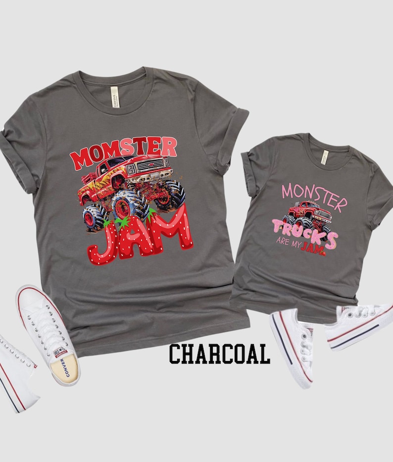 Two charcoal gray t-shirts with monster truck graphics. One shirt says "Momster Jam" with a red monster truck and strawberry design. The other says "Monster Trucks are my Jam" with a red monster truck. White sneakers are on the sides.