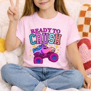 May include: Pink t-shirt with the text "READY TO CRUSH" above a cartoon monster truck with a unicorn head. The truck is purple and pink with blue wheels and a yellow lightning bolt. The shirt is worn by a person.