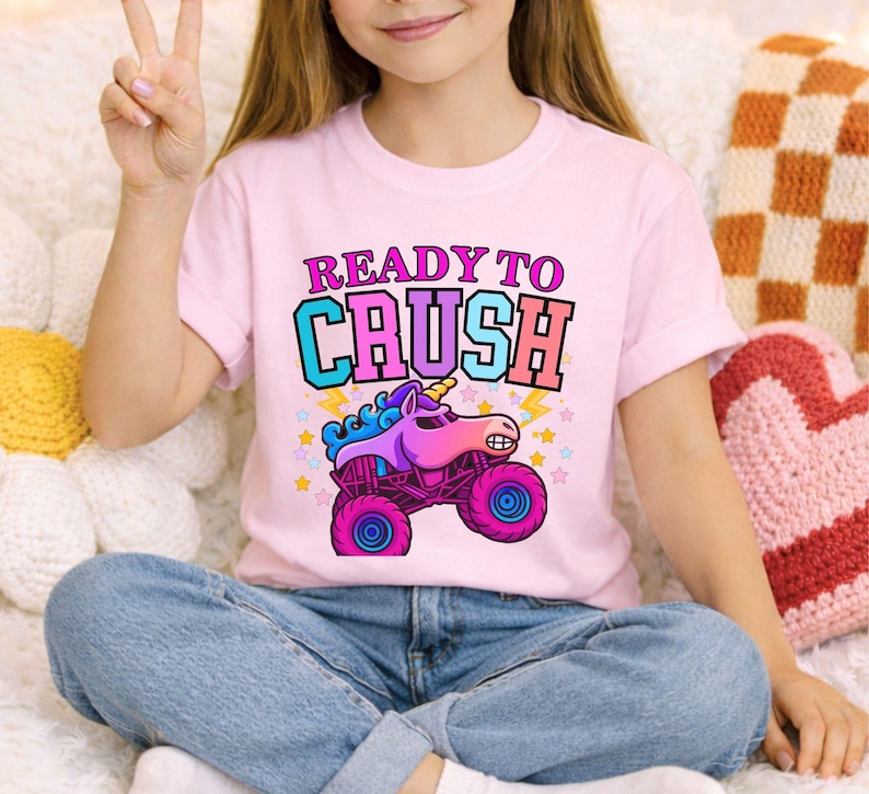 May include: Pink t-shirt with the text "READY TO CRUSH" above a cartoon monster truck with a unicorn head. The truck is purple and pink with blue wheels and a yellow lightning bolt. The shirt is worn by a person.