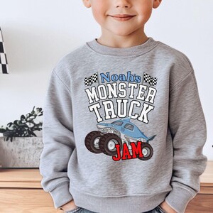 May include: A gray sweatshirt featuring the text "Noah's Monster Truck Jam" with a cartoon shark-themed monster truck design. The design includes black and white checkered flags and red lettering.
