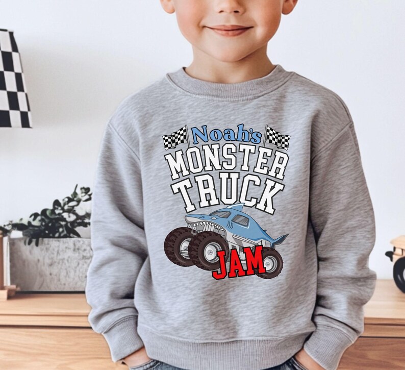 May include: A gray sweatshirt featuring the text "Noah's Monster Truck Jam" with a cartoon shark-themed monster truck design. The design includes black and white checkered flags and red lettering.