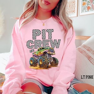 Pictured is a woman wearing a pink sweatshirt with black and white checkered graphic letters that say "PIT CREW" and a cartoon purple and green monster truck.