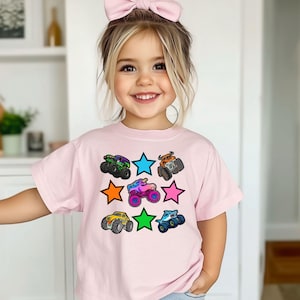 Może przedstawiać: Jasnor&oacute;żowa koszulka z kolorową grafiką monster truck&oacute;w i kształtami gwiazd. Ciężar&oacute;wki są w r&oacute;żnych kolorach, w tym zielonym, pomarańczowym i niebieskim. Koszulka jest przeznaczona dla dzieci.