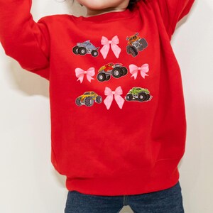 May include: A red sweatshirt featuring colorful monster truck graphics and pink bow accents. The design includes various monster trucks in blue, orange, yellow, and green. The sweatshirt is made of a soft material and is suitable for children.