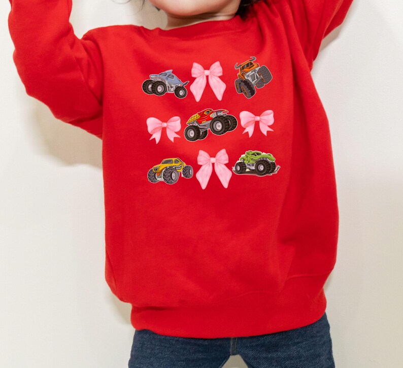 May include: A red sweatshirt featuring colorful monster truck graphics and pink bow accents. The design includes various monster trucks in blue, orange, yellow, and green. The sweatshirt is made of a soft material and is suitable for children.