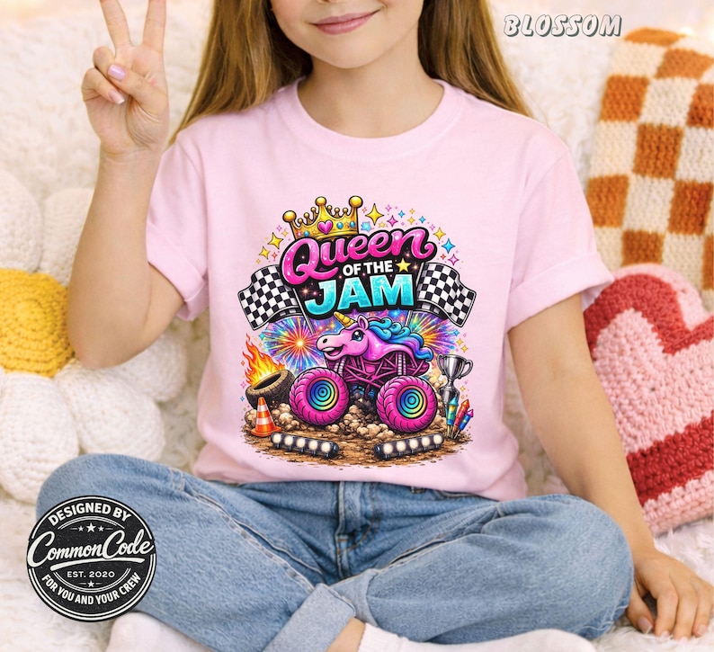 Pictured is a girl wearing a light pink t-shirt featuring a colorful graphic of a unicorn monster truck with the text "Queen of the Jam". The design includes checkered flags, a crown, and a trophy. The shirt is designed by Common Code.