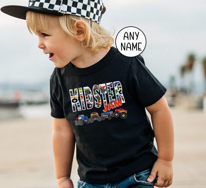 toddler boy model is wearing a black t-shirt with patterned letters that say "Kidster" Jam. The pattern in the letters is colorful monster trucks. Under the word Kidster Jam is a row of 4 multi-colored monster trucks, red, white and blue, green, brown, and orange.