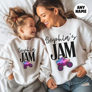 May include: Two white sweatshirts with the text "Sophia's JAM" in black script and block letters. A colorful monster truck graphic is below the text. The sweatshirts are worn by a mother and daughter, lying on a white surface.
