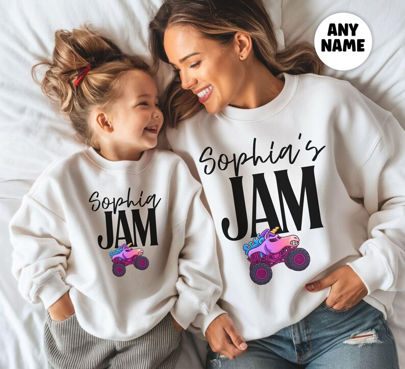 May include: Two white sweatshirts with the text "Sophia's JAM" in black script and block letters. A colorful monster truck graphic is below the text. The sweatshirts are worn by a mother and daughter, lying on a white surface.