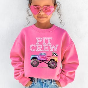 Pictured is a young girl wearing a pink crewneck sweatshirt which says "PIT CREW" in white chalk letters above a pink rainbow colored monster truck with big tires and a checkered flag