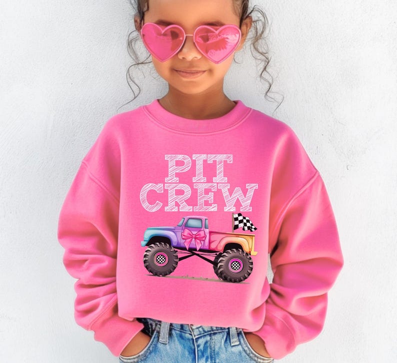 Pictured is a young girl wearing a pink crewneck sweatshirt which says "PIT CREW" in white chalk letters above a pink rainbow colored monster truck with big tires and a checkered flag