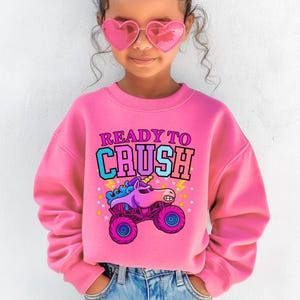 Pu&ograve; includere: Una felpa rosa con la scritta "READY TO CRUSH" sopra un unicorno dei cartoni animati alla guida di un monster truck. La grafica &egrave; nei toni del blu, viola e rosa. La felpa &egrave; a girocollo.