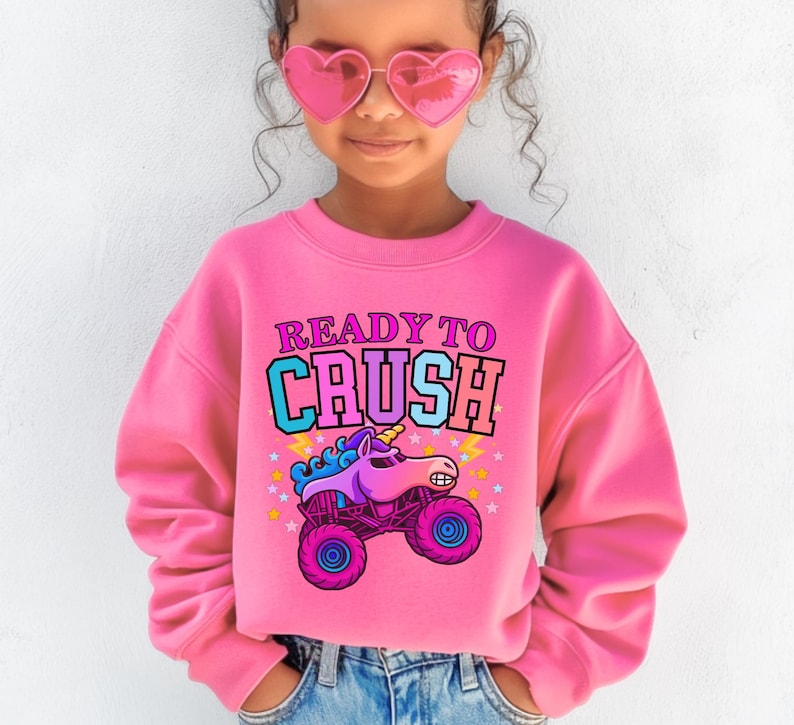 Pu&ograve; includere: Una felpa rosa con la scritta "READY TO CRUSH" sopra un unicorno dei cartoni animati alla guida di un monster truck. La grafica &egrave; nei toni del blu, viola e rosa. La felpa &egrave; a girocollo.