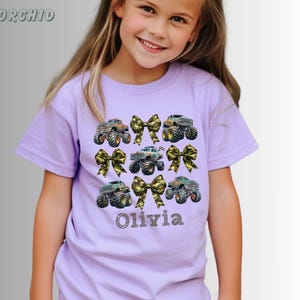 May include: A light purple t-shirt featuring a design of monster trucks and camouflage bows. The name "Olivia" is printed in a script font below the trucks. The shirt is short-sleeved.