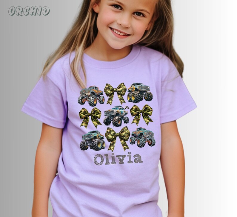 May include: A light purple t-shirt featuring a design of monster trucks and camouflage bows. The name "Olivia" is printed in a script font below the trucks. The shirt is short-sleeved.