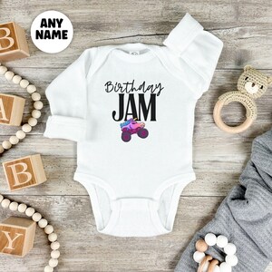 May include: White baby bodysuit with long sleeves. The text "Birthday JAM" is printed in black, with a colorful monster truck graphic. A wooden toy and alphabet blocks are in the background.