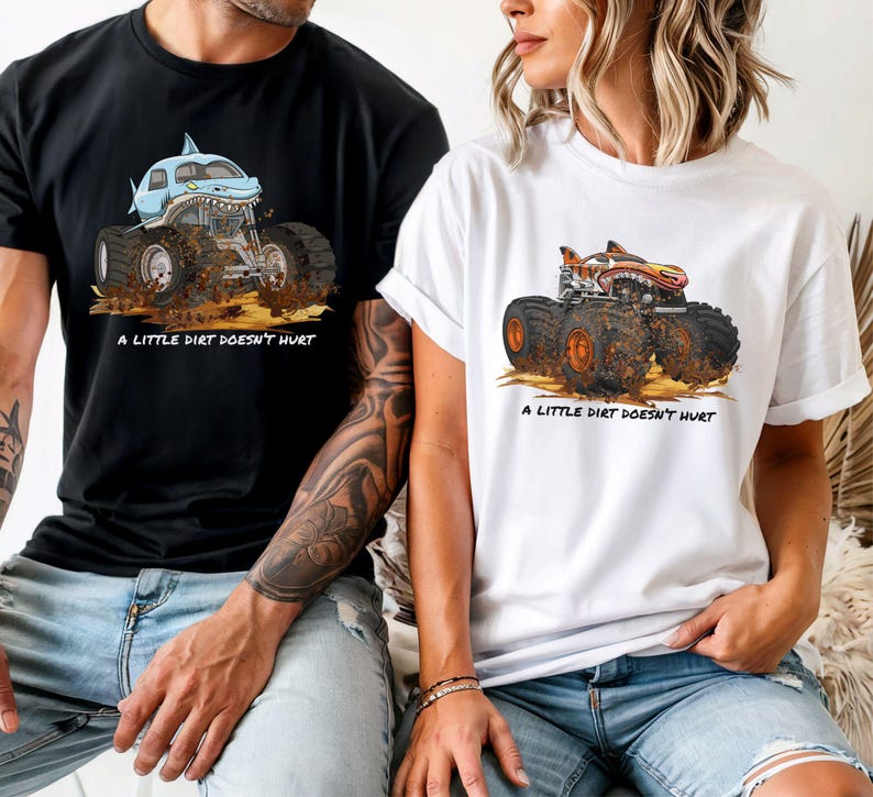 May include: Black and white t-shirts with monster truck graphics. One shirt features a blue shark-themed truck, the other an orange and red truck. Both shirts have the text "A LITTLE DIRT DOESN'T HURT" below the graphic.