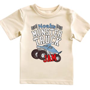 以下が含まれることがあります： クリーム色のTシャツ。大胆なフォントで「Noah's MONSTER TRUCK JAM」と書かれています。デザインには、大きな車輪とチェッカーフラッグを備えたサメ型のモンスタートラックが含まれています。「JAM」という単語は赤色です。